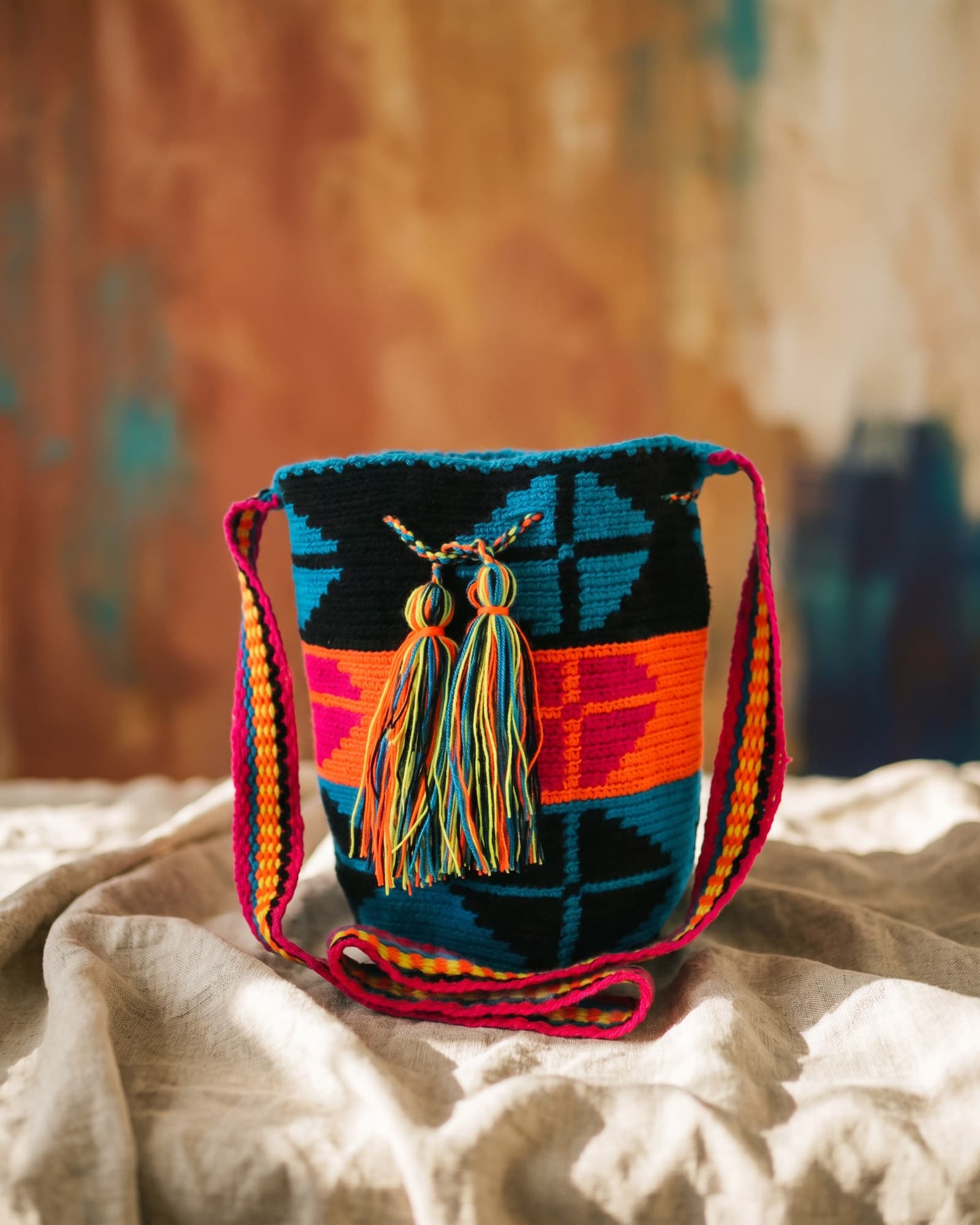 Schwarze Mochila mit dichtem geometrischem Muster in Türkis, Orange und Fuchsia, auf Leinen liegend im warmen Tageslicht.