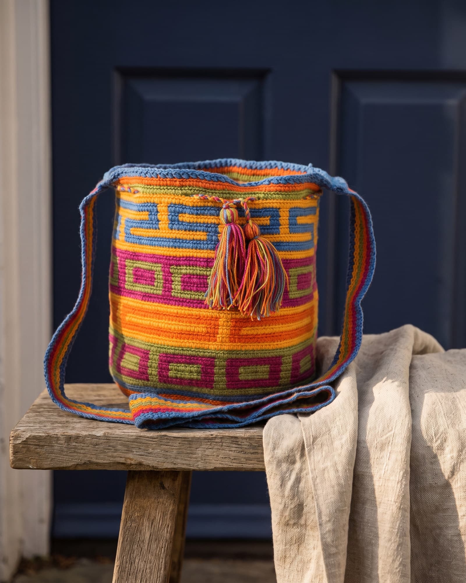 Bunte Mochila mit eckigem Greca-Mäander in Blau, Orange, Magenta und Oliv, auf Leinen liegend im warmen Tageslicht.